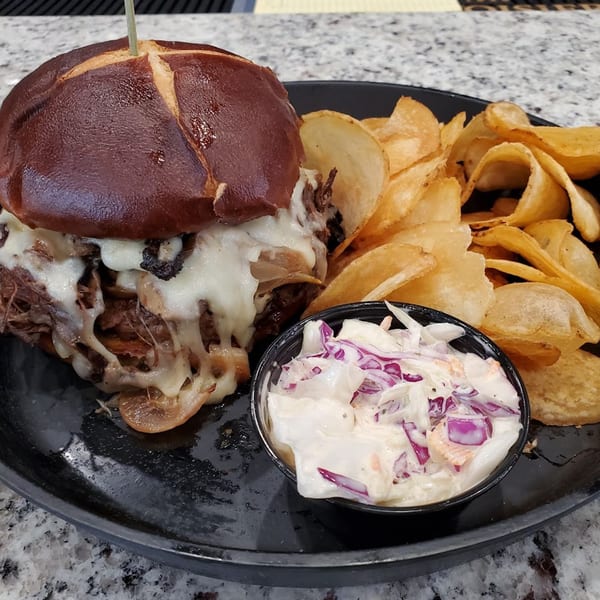 MP Kitchen & Bar's pulled pork sandwich with melted cheese, chips, and coleslaw on a black plate.