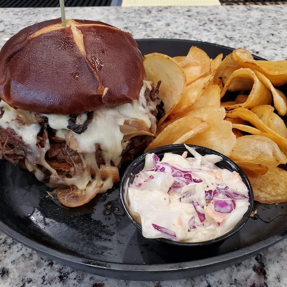 MP Kitchen & Bar's pulled pork sandwich with melted cheese, chips, and coleslaw on a black plate.